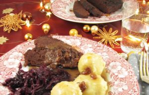 Auf einem Teller liegen zwei Scheiben veganer Sauerbraten, Knödel und Rotkohl. Alles ist mit Sauce übergossen. Im Hintergrund liegt veganer Sauerbraten am Stück auf einer Platte. Darum liegen kleine Christbaumkugeln und Baststerne.