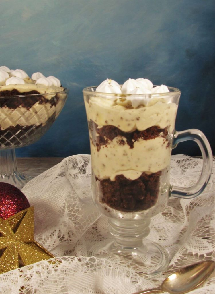 Brownie-Schichtdessert in einem Glas steht auf einer weißen Spitzendecke. Im Hintergrund ist schichtdessert in einer Dessertschale.