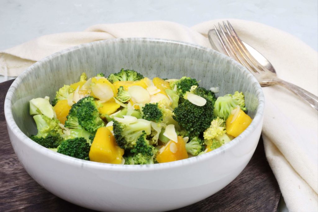 Eine Schale mit Brokkolisalat mit Mango steht auf einem dunkelbraunen Brett. Daneben liegt ein beiges Tuch auf dem silbernes Salatbesteck liegt.