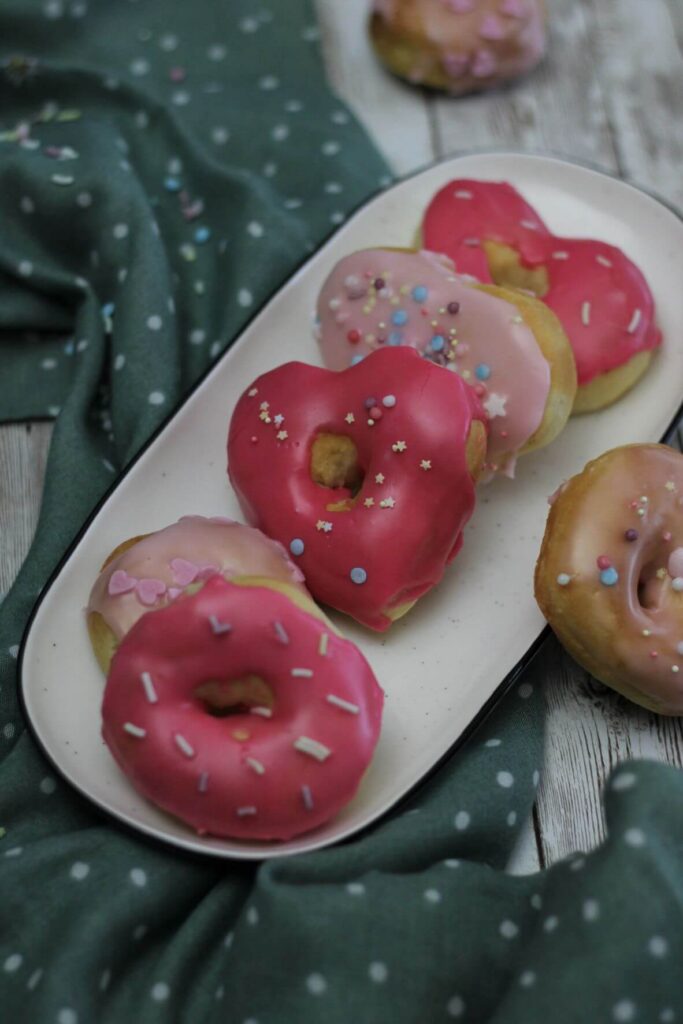 Eine weiße längliche Platte liegt auf einem grüntürkisen Tuch mit weißen Punkten. Auf der Platte liegen mehrere kleine Donuts mit rosa Zuckerguß und bunten Zuckerstreuseln.