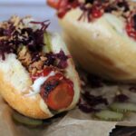 Nahaufnahme von zwei Hot Dogs aus Backpapier. Die Hot Dog Brötchen sind mit einer marinierten Möhre, sauren Gurkenscheiben, Röstzwiebeln, lila Sauerkraut und Ketchup gefüllt.