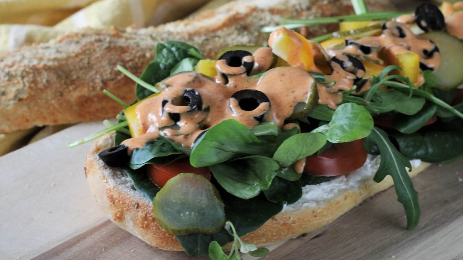 Auf einem Holzbrett liegt ein offenes Subsandwich , dass mit Blattgemüse, gelber Paprika, Tomate und Oliven belegt ist. Auf dem Gemüse sieht man eine orange Sauce.