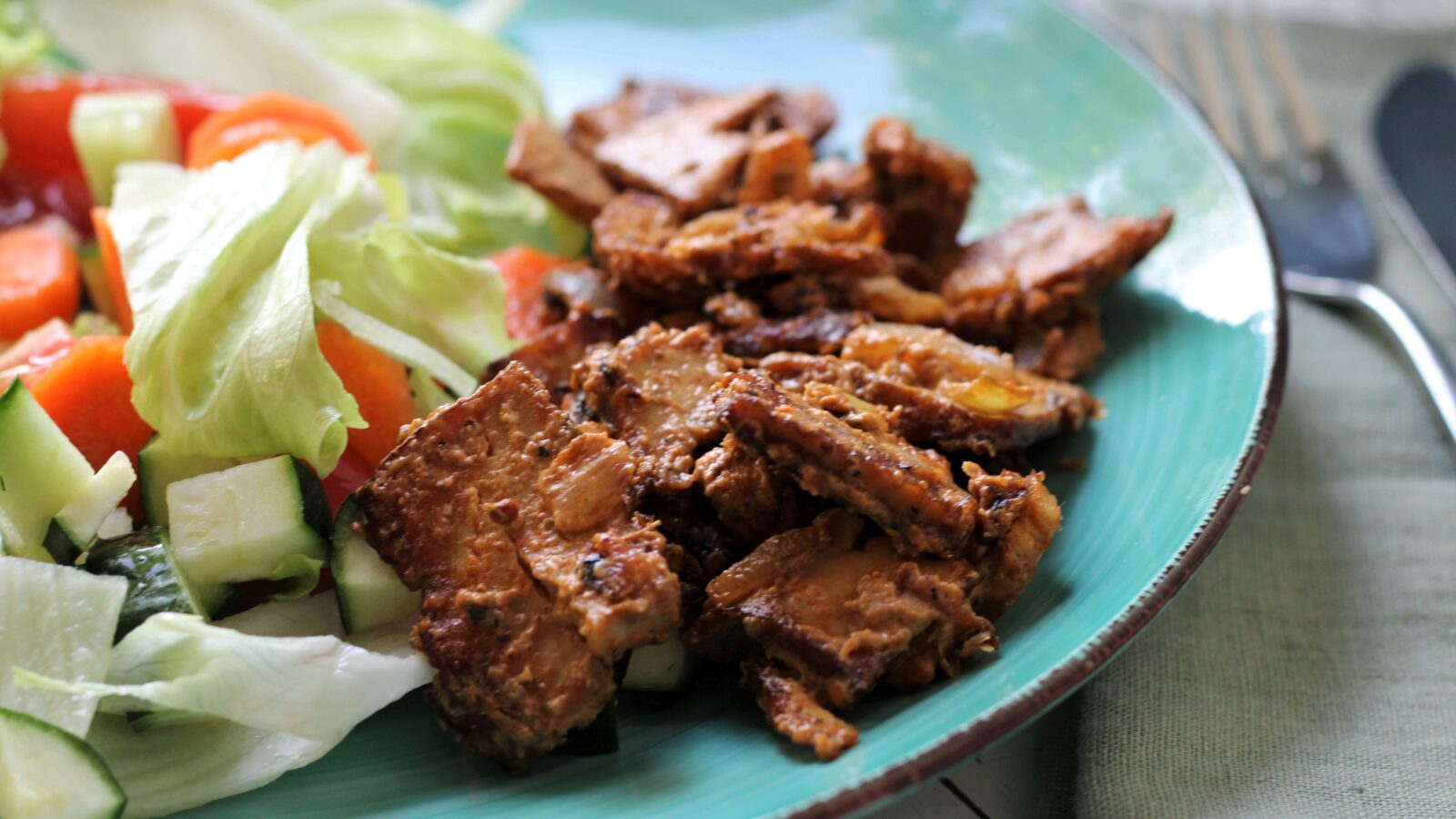 Ein türkiser Teller steht auf einem weißen Holztisch. Daneben liegt eine grünliche Serviette. Auf dem Teller liegt braunes Tofugyros neben gemischtem Salat.