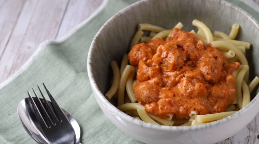 Auf einer weißen Holzplatte liegt eine hellgrüne Stoffserviette. Auf dieser steht ein graues Schälchen, dass mit Nudeln gefüllt ist, die mit einer rötlichen Sauce begossen sind . In der Sauce sind Vleischbällchen erkennbar. Neben der Schüssel liegen ein Löffel und eine Gabel.